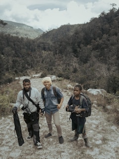 Three people with backpacks stand on a rugged path within a mountainous and forested landscape. The individuals appear to be prepared for an outdoor adventure or trek, carrying equipment such as a camera and tripod.