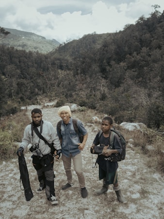 Three people with backpacks stand on a rugged path within a mountainous and forested landscape. The individuals appear to be prepared for an outdoor adventure or trek, carrying equipment such as a camera and tripod.
