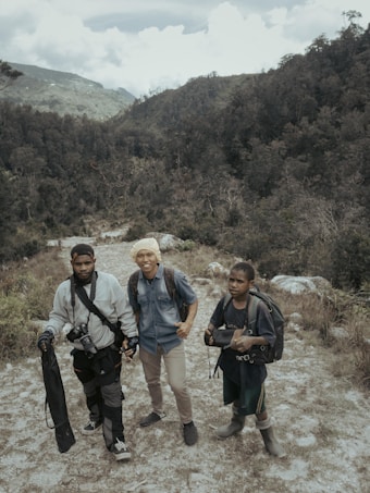 Three people with backpacks stand on a rugged path within a mountainous and forested landscape. The individuals appear to be prepared for an outdoor adventure or trek, carrying equipment such as a camera and tripod.