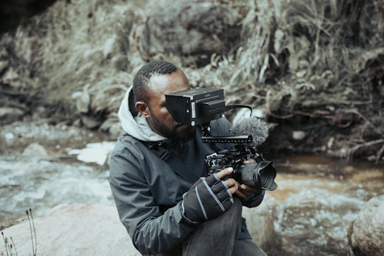 A person is wearing warm clothing and gloves, holding a professional camera setup with additional equipment, while sitting near a river with rocky surroundings and dried vegetation.