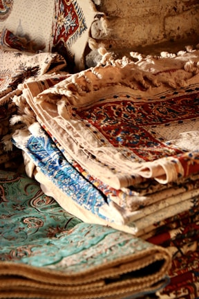A stack of intricately woven textiles or carpets, displaying a variety of patterns and colors. The fabrics have detailed floral and geometric designs in shades of red, blue, and beige, laid over one another, creating a rich tapestry of textures.