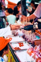 A street food vendor wearing a glove hands a skewer of candied fruit to a customer over a food stall counter. The counter is lined with trays of glossy, red candied fruits on skewers. Children and patrons are gathered around, creating a busy, lively atmosphere.