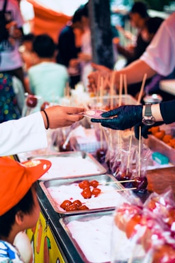 A street food vendor wearing a glove hands a skewer of candied fruit to a customer over a food stall counter. The counter is lined with trays of glossy, red candied fruits on skewers. Children and patrons are gathered around, creating a busy, lively atmosphere.