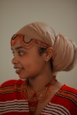 A person wearing a tan headwrap and colorful beaded jewelry, including a necklace and headpiece. The clothing features a red and black pattern.