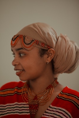 A person wearing a tan headwrap and colorful beaded jewelry, including a necklace and headpiece. The clothing features a red and black pattern.