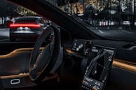 The interior of a modern car with a sleek design, featuring a steering wheel and two digital screens displaying navigation and car information. A vehicle is visible through the windshield, partially lit by red taillights. The ambient lighting creates a futuristic and high-tech atmosphere.