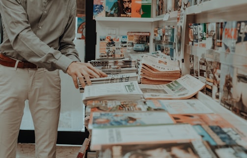 A person is browsing through a magazine stand or newsstand. Numerous magazines and newspapers are neatly arranged on the shelves and counter. The assortment includes various titles and covers, featuring different colors and images. The person is wearing a light-colored shirt and pants and is positioned with their hands hovering over some of the publications.