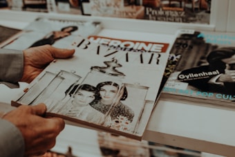 A person is holding several magazines, with the focus on a vintage-style magazine cover featuring two women in black and white. Other magazines are stacked on the table, and the surrounding area is softly focused.