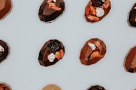 A collection of geometrically shaped chocolates is arranged on a light surface. Each piece is embellished with a variety of toppings, including nuts, dried fruits, and small white pieces.