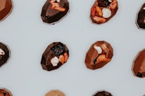 A collection of geometrically shaped chocolates is arranged on a light surface. Each piece is embellished with a variety of toppings, including nuts, dried fruits, and small white pieces.