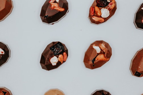 A collection of geometrically shaped chocolates is arranged on a light surface. Each piece is embellished with a variety of toppings, including nuts, dried fruits, and small white pieces.