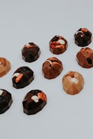A collection of geometric-shaped chocolates arranged on a light surface. The chocolates have various fillings, including nuts and dried fruits, creating a visually appealing assortment.
