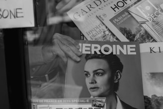 A black and white image of a person’s hand holding a magazine titled 'HEROINE' with a portrait of a woman on the cover. The background shows various other magazines and newspapers stacked together.