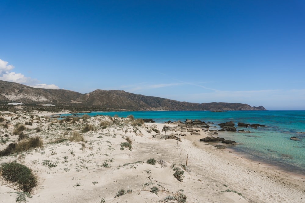 a sandy beach with blue water and mountains in the background