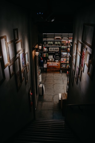 A dimly lit staircase leads downward to a room filled with shelves stocked with board games. The walls lining the staircase are adorned with empty picture frames, and faint lighting comes from wall-mounted lamps. There are boxes at the bottom of the stairs, and the space has a cozy, intimate feeling.