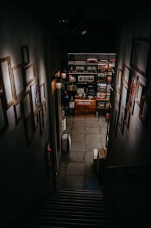 A dimly lit staircase leads downward to a room filled with shelves stocked with board games. The walls lining the staircase are adorned with empty picture frames, and faint lighting comes from wall-mounted lamps. There are boxes at the bottom of the stairs, and the space has a cozy, intimate feeling.