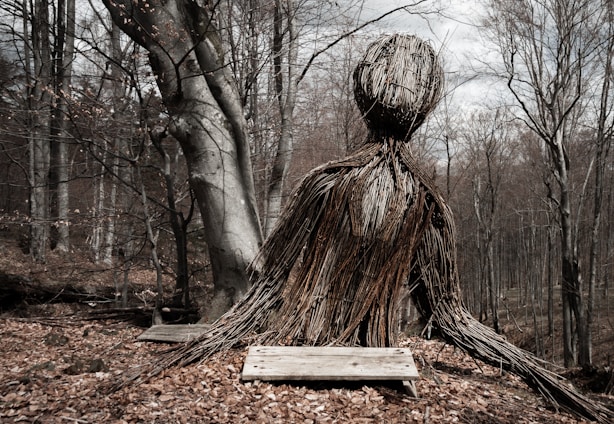 A large sculpture made of intertwined twigs and branches shaped like a humanoid figure sits in a forest. The forest is composed of bare trees, indicative of late autumn or winter, with a ground covered in fallen leaves. The sculpture appears to be seated on wooden planks, and the sky is partly cloudy.