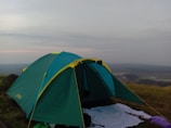 A green and yellow camping tent set up on a grassy hilltop with a vast landscape in the background. The sky appears cloudy with hints of sunlight breaking through. A white sheet and some camping gear are placed outside the tent.