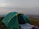 A green and yellow camping tent set up on a grassy hilltop with a vast landscape in the background. The sky appears cloudy with hints of sunlight breaking through. A white sheet and some camping gear are placed outside the tent.
