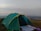 A green and yellow camping tent set up on a grassy hilltop with a vast landscape in the background. The sky appears cloudy with hints of sunlight breaking through. A white sheet and some camping gear are placed outside the tent.