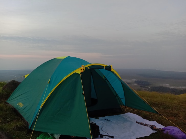 A green and yellow camping tent set up on a grassy hilltop with a vast landscape in the background. The sky appears cloudy with hints of sunlight breaking through. A white sheet and some camping gear are placed outside the tent.