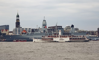 A large naval ship is docked near an urban waterfront with a distinctive clock tower in the background. In front of the naval vessel, a vintage steamboat with a large crowd on deck passes by on the river. The setting is industrial and city-like with modern buildings visible in the distance.