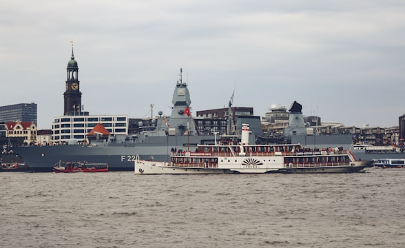 A large naval ship is docked near an urban waterfront with a distinctive clock tower in the background. In front of the naval vessel, a vintage steamboat with a large crowd on deck passes by on the river. The setting is industrial and city-like with modern buildings visible in the distance.