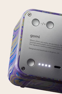 A close-up view of a device featuring a smooth metallic surface with buttons and text. The casing has a colorful marbled pattern. The text includes branding and a message about using recycled materials.