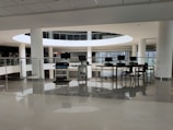 A spacious library interior featuring a modern design with large circular openings and prominent use of white surfaces. Several computers are arranged on a table with chairs, situated near glass railings that overlook a lower level filled with shelves of books. The environment appears clean and well-lit with natural light coming through large windows.