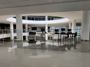 A spacious library interior featuring a modern design with large circular openings and prominent use of white surfaces. Several computers are arranged on a table with chairs, situated near glass railings that overlook a lower level filled with shelves of books. The environment appears clean and well-lit with natural light coming through large windows.