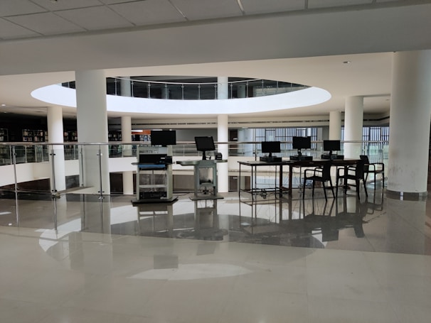 A spacious library interior featuring a modern design with large circular openings and prominent use of white surfaces. Several computers are arranged on a table with chairs, situated near glass railings that overlook a lower level filled with shelves of books. The environment appears clean and well-lit with natural light coming through large windows.