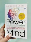 A hand is holding a book titled 'The Power of Your Subconscious Mind' by Dr. Joseph Murphy. The cover features a silhouette of a head with a colorful array of birds flying away from it, symbolizing thoughts or ideas. There is a yellow circle indicating 'Millions of Copies Sold.'