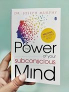 A hand is holding a book titled 'The Power of Your Subconscious Mind' by Dr. Joseph Murphy. The cover features a silhouette of a head with a colorful array of birds flying away from it, symbolizing thoughts or ideas. There is a yellow circle indicating 'Millions of Copies Sold.'
