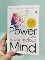 A hand is holding a book titled 'The Power of Your Subconscious Mind' by Dr. Joseph Murphy. The cover features a silhouette of a head with a colorful array of birds flying away from it, symbolizing thoughts or ideas. There is a yellow circle indicating 'Millions of Copies Sold.'