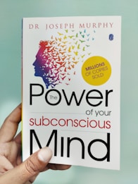 A hand is holding a book titled 'The Power of Your Subconscious Mind' by Dr. Joseph Murphy. The cover features a silhouette of a head with a colorful array of birds flying away from it, symbolizing thoughts or ideas. There is a yellow circle indicating 'Millions of Copies Sold.'