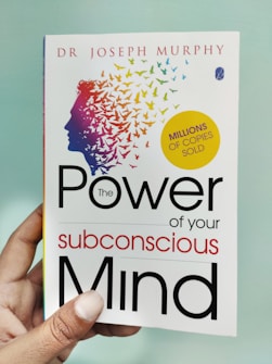 A hand is holding a book titled 'The Power of Your Subconscious Mind' by Dr. Joseph Murphy. The cover features a silhouette of a head with a colorful array of birds flying away from it, symbolizing thoughts or ideas. There is a yellow circle indicating 'Millions of Copies Sold.'