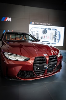 A red BMW M4 car is prominently displayed in a showroom. The car's front grill and headlights are visible, alongside a showroom display in the background highlighting 'Performance Parts'. The interior, with leather seats, is partially visible through the windshield.