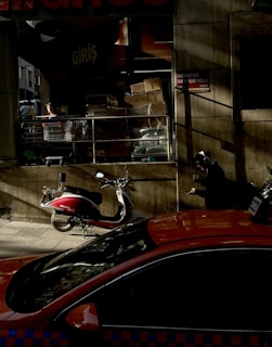 A street scene featuring a storefront with stacked cardboard boxes near the entrance. A person is sitting on the sidewalk, engrossed in their phone, wearing headphones. A bright red motorbike and a red vehicle, possibly a taxi, are parked nearby, casting shadows on the pavement.