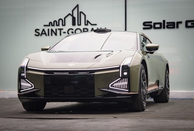 A sleek, modern car with a futuristic design is parked indoors. The vehicle features distinctive LED headlights and a reflective, streamlined body. The background includes a logo on a glass wall, suggesting an upscale or showroom environment.