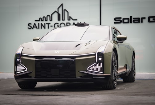 A sleek, modern car with a futuristic design is parked indoors. The vehicle features distinctive LED headlights and a reflective, streamlined body. The background includes a logo on a glass wall, suggesting an upscale or showroom environment.