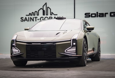 A sleek, modern car with a futuristic design is parked indoors. The vehicle features distinctive LED headlights and a reflective, streamlined body. The background includes a logo on a glass wall, suggesting an upscale or showroom environment.