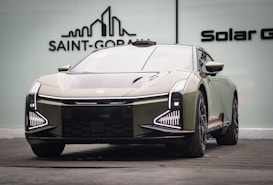 A sleek, modern car with a futuristic design is parked indoors. The vehicle features distinctive LED headlights and a reflective, streamlined body. The background includes a logo on a glass wall, suggesting an upscale or showroom environment.