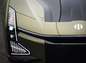A close-up view of the front end of a modern vehicle, showcasing sleek, angular design elements and LED headlights. The car has a futuristic look with a glossy finish and a distinct emblem on the hood.