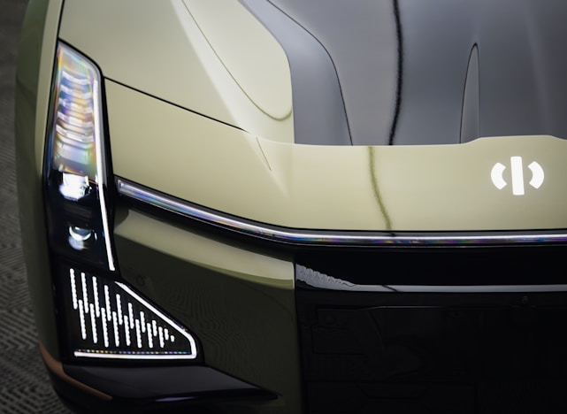 A close-up view of the front end of a modern vehicle, showcasing sleek, angular design elements and LED headlights. The car has a futuristic look with a glossy finish and a distinct emblem on the hood.