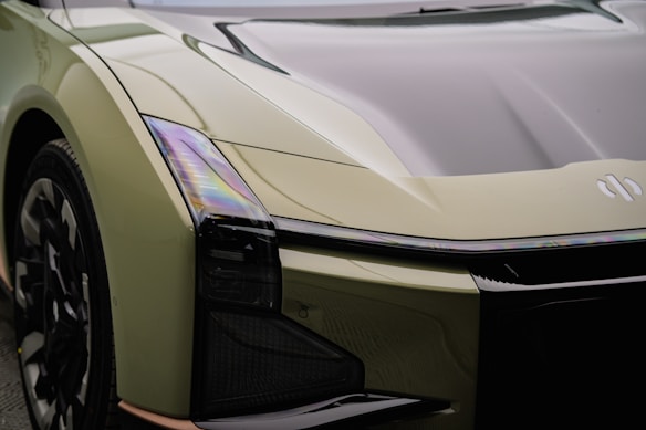 A futuristic car featuring sleek, angular lines and a glossy surface. The vehicle has a distinct headlamp design and a reflective hood. The paint appears to be a light olive green with iridescent reflections.