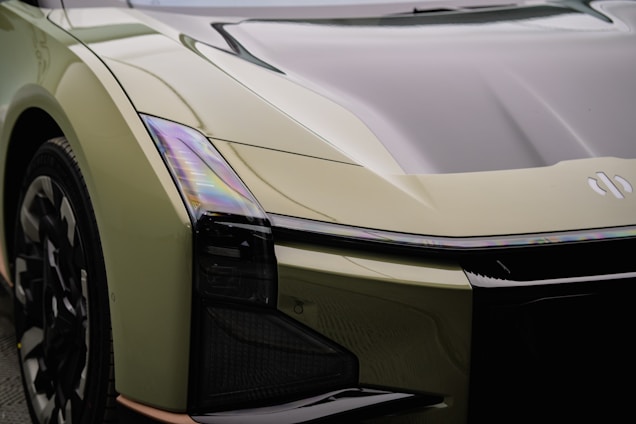 A futuristic car featuring sleek, angular lines and a glossy surface. The vehicle has a distinct headlamp design and a reflective hood. The paint appears to be a light olive green with iridescent reflections.