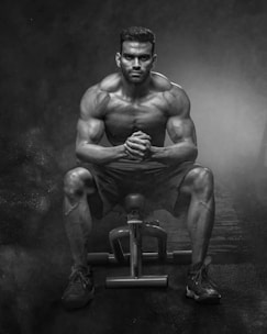 A muscular man is sitting on a workout bench in a gym environment, displaying a focused and intense expression. The lighting casts strong shadows, highlighting his defined muscles and creating a dramatic atmosphere.