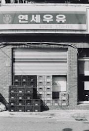A stack of plastic crates is placed in front of a closed metal shutter of a storefront. The building is made of brick, and a sign with Korean text is mounted above.
