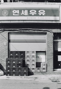 A stack of plastic crates is placed in front of a closed metal shutter of a storefront. The building is made of brick, and a sign with Korean text is mounted above.