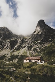 A rustic mountain lodge nestled among lush vegetation with towering rocky mountains in the background. The sky is partly cloudy, casting soft shadows over the landscape. A small body of water lies in the foreground, reflecting the serene environment.